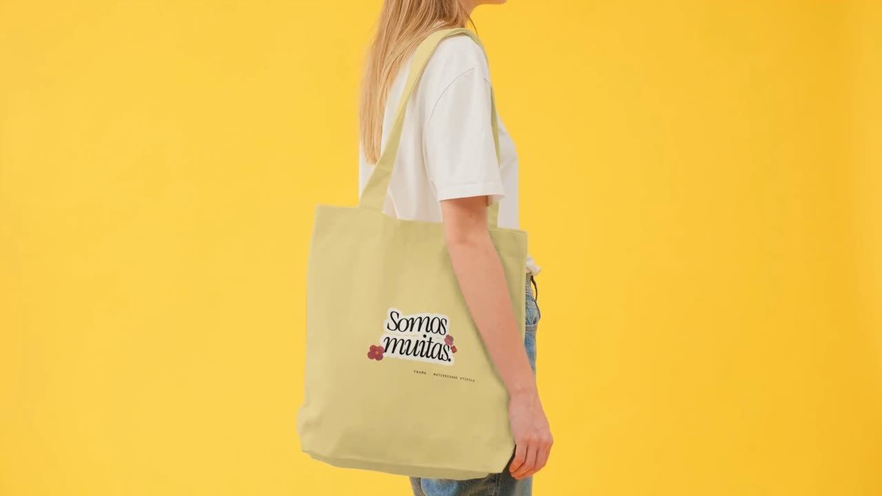 Mulher de camiseta branca carrega uma ecobag com o adesivo Somos muitas, sobre fundo amarelo